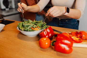 Close up of young woman preparing fresh salad in a bowl, mixing ingredients with forks