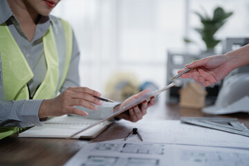 Two engineers are discussing building plans using a tablet and blueprints, collaborating on a...
