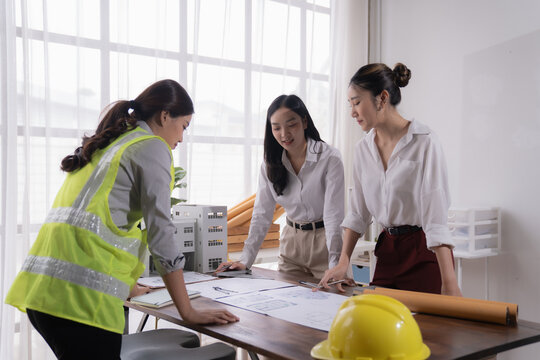 Three asian female engineers and architects are working together on a project using blueprint and a scale model of a building in a bright office