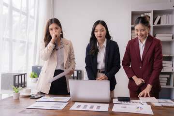 Three asian businesswomen are working together, analyzing financial data using a laptop and documents spread out on a desk in a modern office meeting, showing teamwork and collaboration