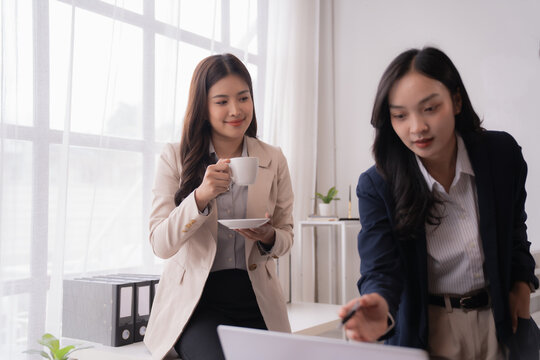 Two young businesswomen are discussing a project, with one pointing at a laptop and the other drinking coffee, showcasing teamwork and collaboration in a modern office environment
