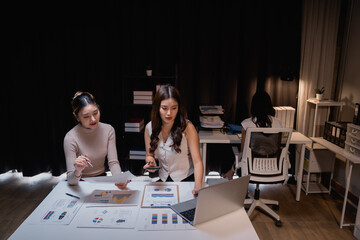 Two asian businesswomen are working late at night in the office, using a laptop and analyzing financial charts and documents spread out on the table, while another colleague works in the background
