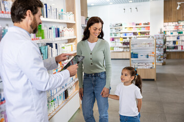 Obraz premium Young mom with a cute kid choosing medicine in the pharmacy, male pharmacist helping her