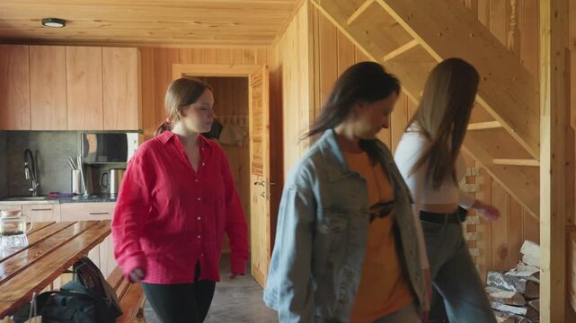 three young women inside wooden cabin kitchen look toward staircase as one climbs up, others observe with interest, natural daylight brightens rustic space with timber walls, visible logs