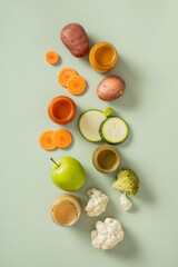 Vertical flat lay of fresh vegetables, fruits, and colorful purees in jars on pastel green background. Baby food, healthy eating, and organic nutrition concept.