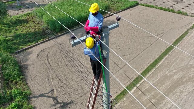 Bogura, Bangladesh - 21 August 2025: Aerial view of electrical workers in yellow hardhats, atop a ladder, repairing a towering utility pole in a rural landscape.