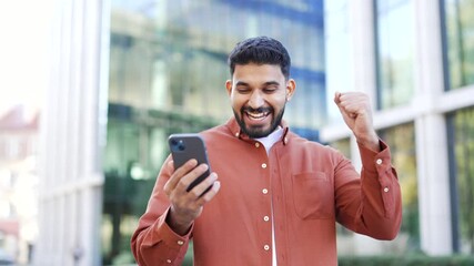 Satisfied happy young businessman received great news on mobile phone while standing on street near modern office building. Smiling joyful handsome man reads a positive good message on a smartphone - Powered by Adobe