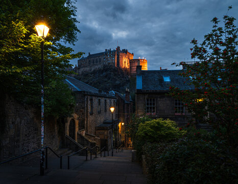 Blick vom Vennel Viewpoint in Edinburgh zur blauen Stunde auf Edinburgh Castle, gelb beleuchtete Burg auf dem Castle Rock, Fackeln auf den Zinnen und gelbe Stra&szlig;enlaternen entlang der Gasse