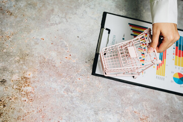 Hand Placing Mini Shopping Cart on Clipboard with Business Graphs and Charts Background