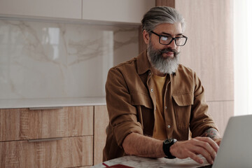 Horizontal medium portrait shot of handsome mature man with beard on face wearing eyeglasses sitting at table surfing Internet on laptop