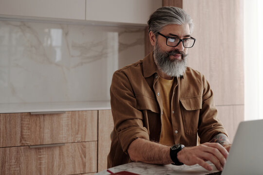 Horizontal medium portrait shot of handsome mature man with beard on face wearing eyeglasses sitting at table surfing Internet on laptop
