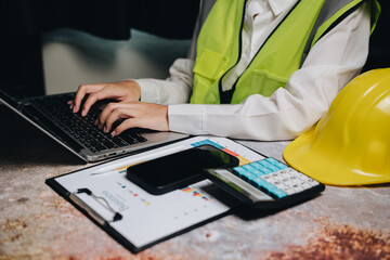 Business professional in safety vest working on laptop with calculator, smartphone, and clipboard on desk