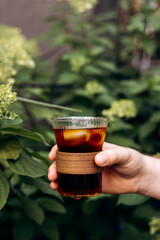Hand holding a glass of cold brew coffee with ice on green outdoor table.