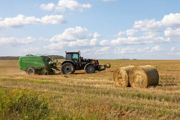 Obraz premium Round hay bales in field with baler wrapper machine working in background