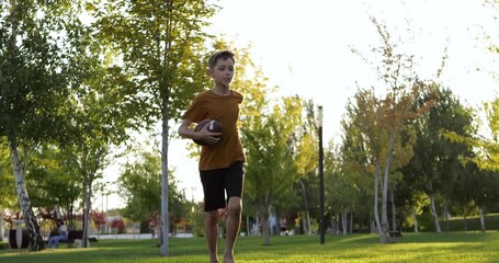 Teen boy runs with rugby ball on green lawn at sunset during outdoor training. Youth rugby action shows energy, focus, fitness, healthy lifestyle in summer park.Teenager rugby action highlights energy - Powered by Adobe