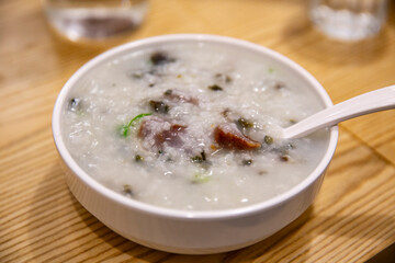 Hot bowl of Cantonese style congee served in white ceramic bowl with Chinese sausage and pickled vegetables on wooden table background