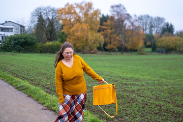 Happy Mature woman in mustard sweater and plaid skirt walking outdoors, holding yellow bag, autumn...