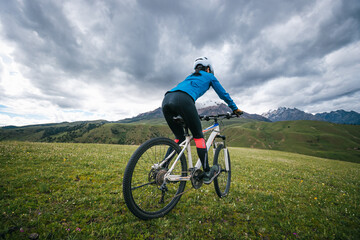 Fototapeta premium Woman cyclist riding mountain bike on grassland mountains with snow capped mountain in the distance