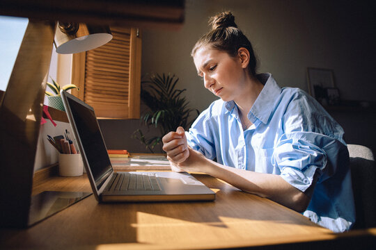 A tired young woman in casual clothes is holding her wrist. The girl is experiencing pain in her arm and is sitting in front of her laptop.