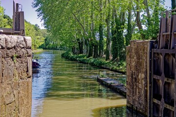 Canal with lush green trees lining the waterway and stone lock gates in Canal du Midi, France.