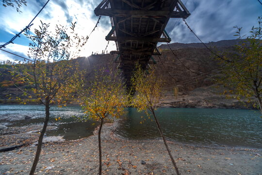 View of a suspension bridge casting shadows on the riverbank, framed by autumn trees against a backdrop of rugged mountains, Yasin Valley, Gilgit Baltistan, Pakistan.