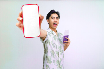 Portrait of young asian man in summer shirt holding passport and ticket while showing large smartphone mockup screen with happy face, concept travel and mobile advertisement.