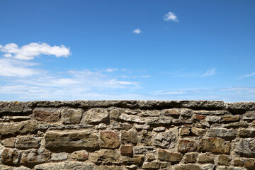 Weathered masonry stone wall against a bright blue sky with light clouds; rustic background and texture with large copy space for design.
