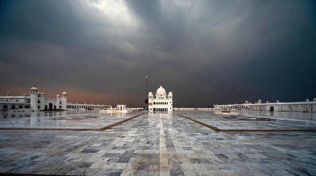 View of a gleaming white Gurdwara complex reflecting the dramatic, stormy sky on a vast marble courtyard, Kartarpur, Punjab, Pakistan.