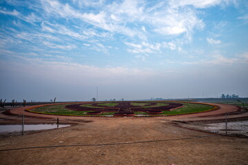 View of an expansive landscape where a large symbol is centered within a circular green space surrounded by a dirt path, under a sky streaked with clouds, Kartarpur, Punjab, Pakistan.