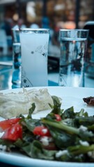 Image of a dining table with a white plate filled with food, Turkish raki including a creamy dish and a salad with tomatoes and greens.