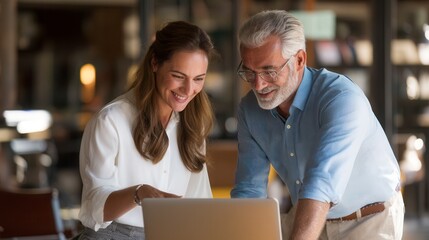 Happy caucasian female adult professional and senior male colleague smiling while looking at laptop together in modern office space, intergenerational business mentoring