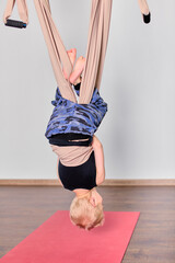 Blonde boy hangs upside down on beige aerial hammock in studio with wooden floor. Wearing blue and...