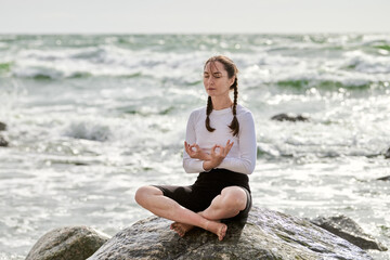 Young woman with braided hair meditates on rocky shore, eyes closed in focus. Sunrise casts warm...