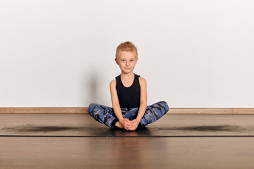 Young blond boy performs yoga in serene studio, seated cross-legged on mat. Light wood floor and plain background create peaceful, focused atmosphere. Natural lighting enhances tranquility