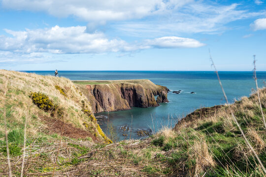 View of rugged cliffs cascading into the turquoise sea under a vast sky spotted with clouds, the land adorned with green grass, Stonehaven, Scotland, United Kingdom.
