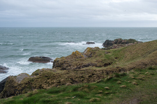 View of rugged, moss-covered cliffs meet the turbulent, grey-blue sea under a cloudy sky, a serene yet wild panorama, Peterhead, Scotland, United Kingdom.