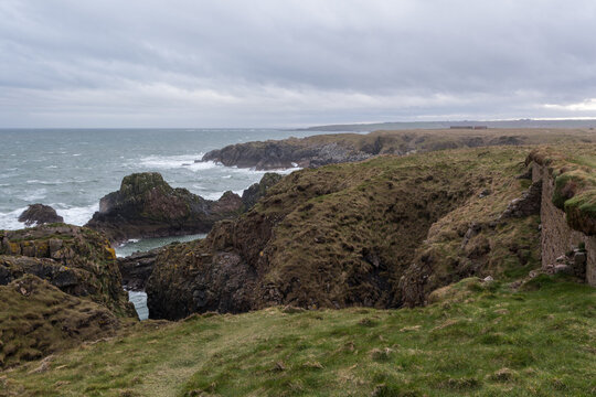 View of rugged cliffs meet the turbulent sea under a brooding sky, with the ruins of a stone structure standing guard, Peterhead, Scotland, United Kingdom.