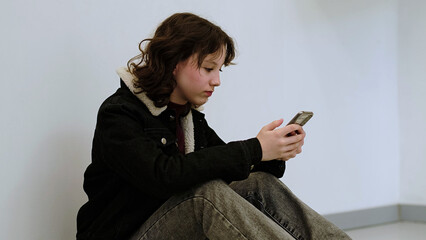 A child in casual attire uses her smartphone while sitting against a blank wall. The photo highlights the pervasive role of technology in everyday life and youth s engagement with digital devices.
