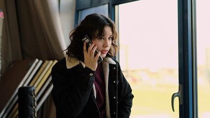 A calm young girl makes or receives a phone call while standing near a window. This image portrays themes of communication, youth, and the importance of staying connected.