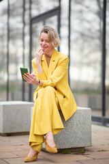Fototapeta premium Blonde woman in a bright yellow pantsuit sitting on a stone bench, smiling while looking at her mobile phone, enjoying a casual moment outside