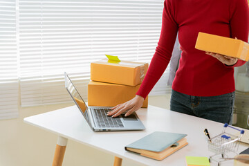 Asian woman in apron using laptop and receipt box and checking online orders to prepare parcels for...