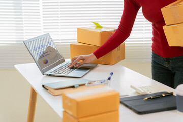 Asian woman in apron using laptop and receipt box and checking online orders to prepare parcels for...