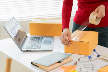 Asian woman in apron using laptop and receipt box and checking online orders to prepare parcels for...