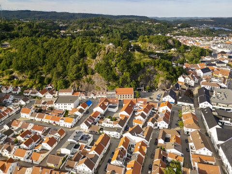 Aerial view of the town nestled between the rugged cliffs and the sea, a tapestry of red roofs under the soft sky, Mandal, Agder, Norway.