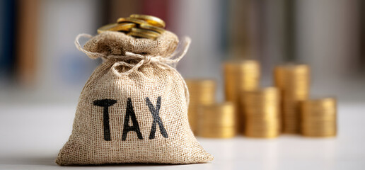 burlap sack with the word tax filled with gold coins placed on white table blurred stacks of coins in background business concept symbolizing banking financial security investment growth and economic