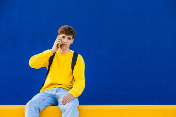 Young man wearing yellow sweatshirt and backpack talking on smartphone in front of blue wall
