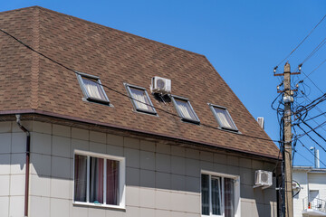 Residential building featuring textured brown shingle roof, multiple angled skylight windows, and exposed air conditioner unit, alongside complex utility pole with numerous power lines under bright bl