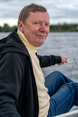 Genuine portrait of smiling mature man with blue eyes, wearing cream turtleneck and black hooded jacket, sitting on boat with calm water and distant shore under overcast sky.