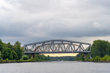 Obraz premium Grand steel truss railway bridge gracefully spans wide, serene river or canal, bordered by dense green forests and distant boats navigating under overcast sky.