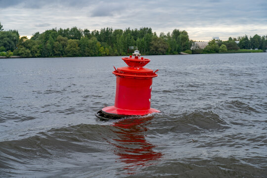 Vibrant red navigational buoy floating prominently in tranquil river with gentle ripples, set against lush green treelined shore under overcast sky